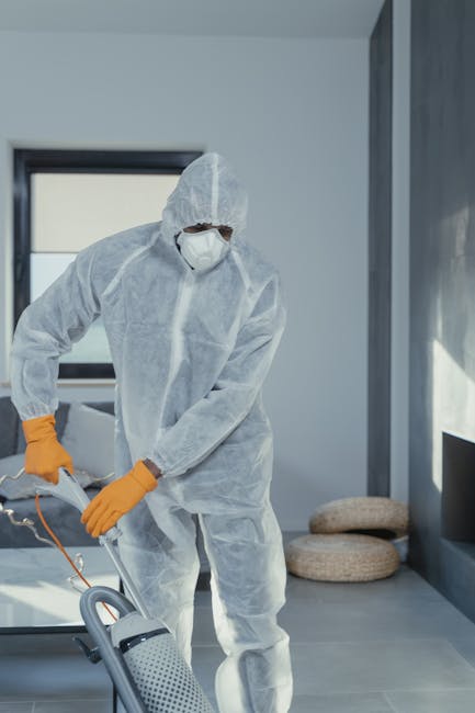 A professional cleaner dressed in a white protective suit, mask, and bright orange gloves is operating a vacuum cleaner on a smooth, gray tiled floor in a modern, well-lit room with large windows allo