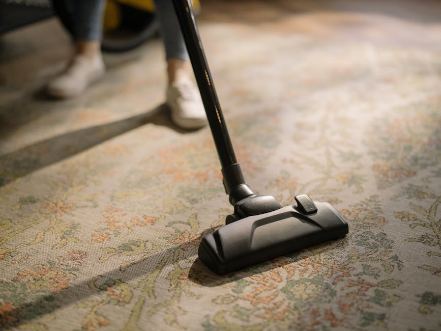 A professional cleaner dressed in a white protective suit, mask, and bright orange gloves is operating a vacuum cleaner on a smooth, gray tiled floor in a modern, well-lit room with large windows allo